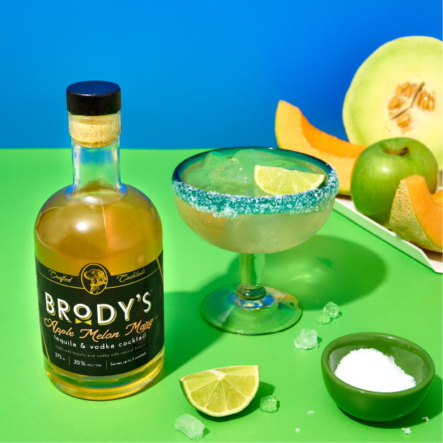 Bottle of Brody's Apple Melon Margarita with a cocktail glass, lime, and melon on a colorful background