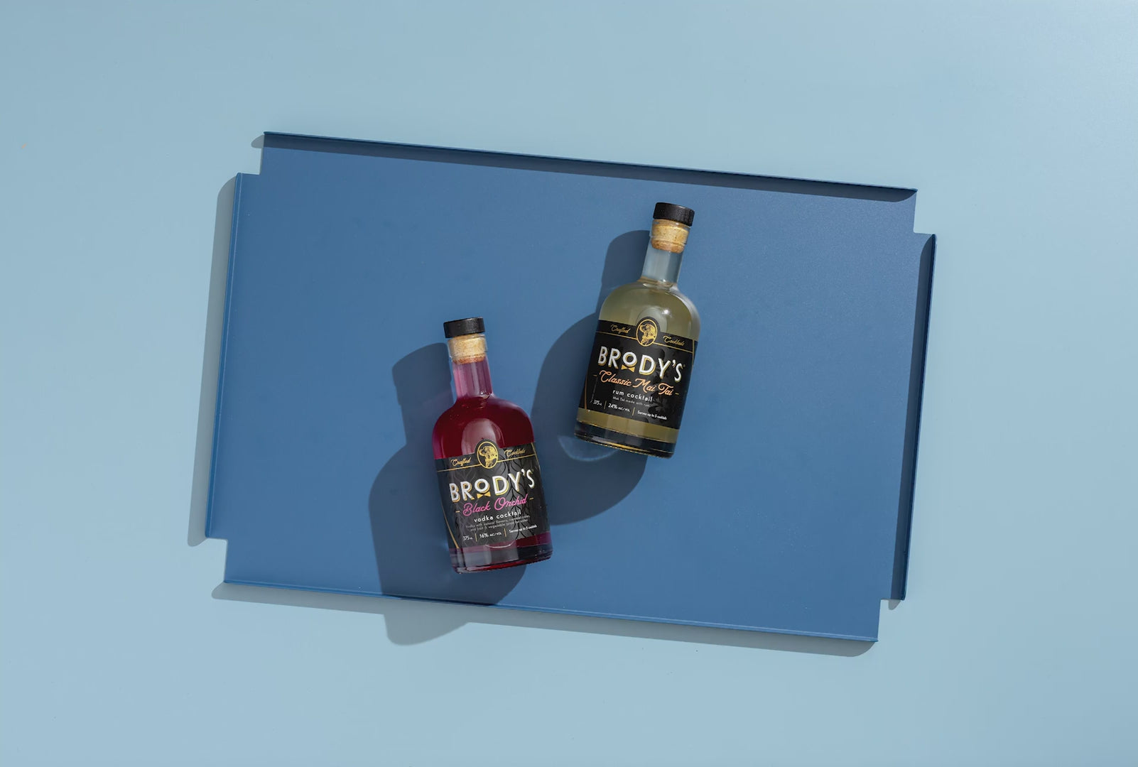 Brody’s Crafted Cocktails Classic Mai Tai and Black Orchid bottles on a minimal blue background, showcasing premium ready-to-serve cocktails with the slogan “Sophisticated Taste. Simply Served.”